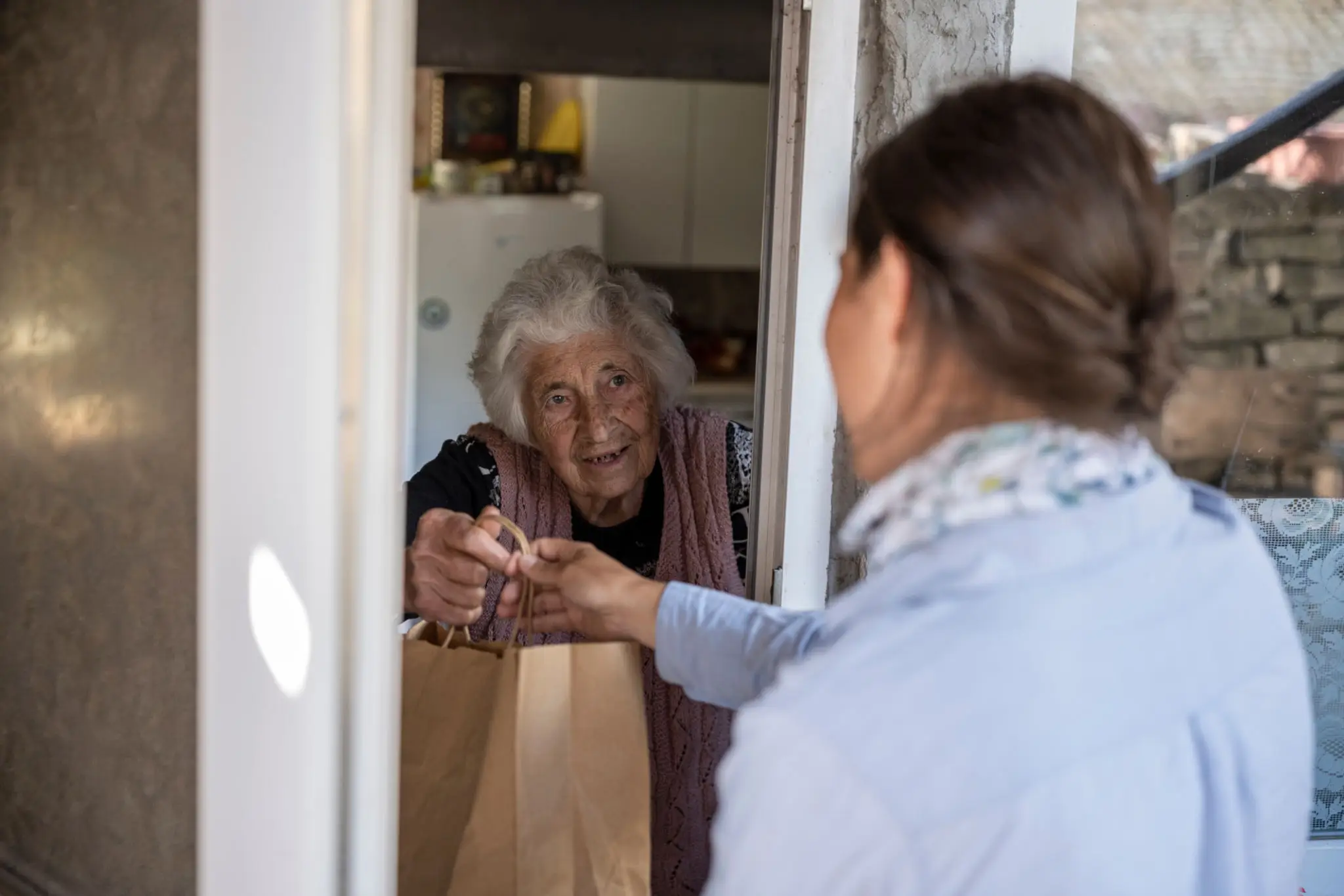 A woman hands a paper bag to an elderly woman through a door.