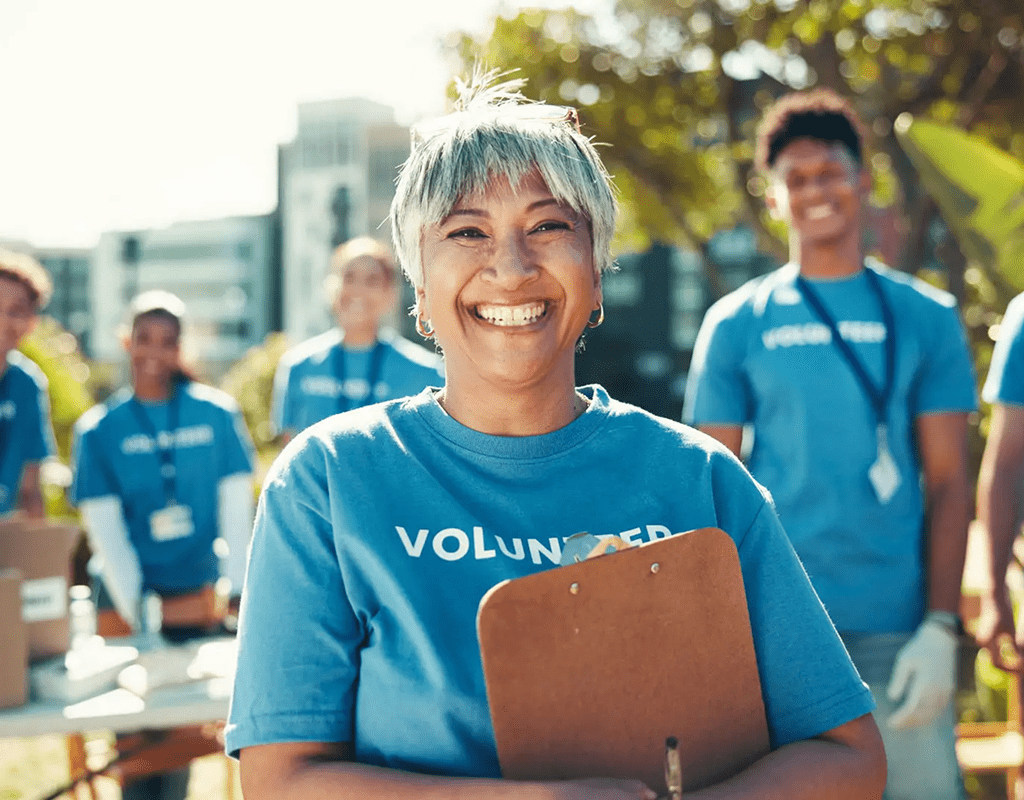 Smiling volunteer holding a clipboard outdoors with a team behind her.