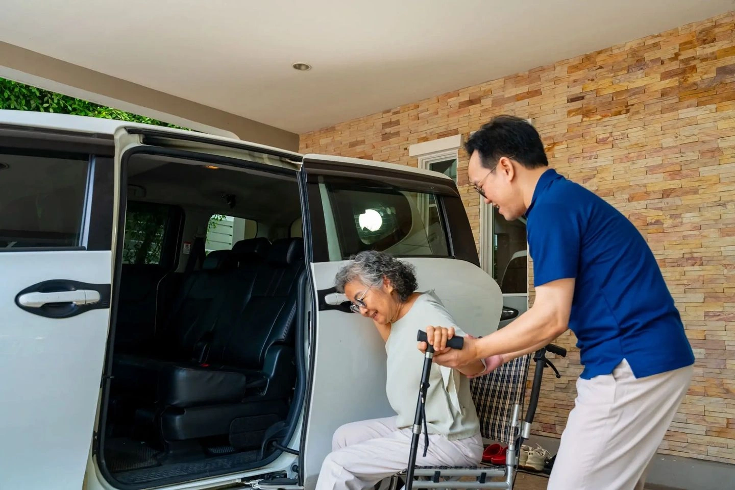 A man assists an elderly woman into a vehicle using a walker.