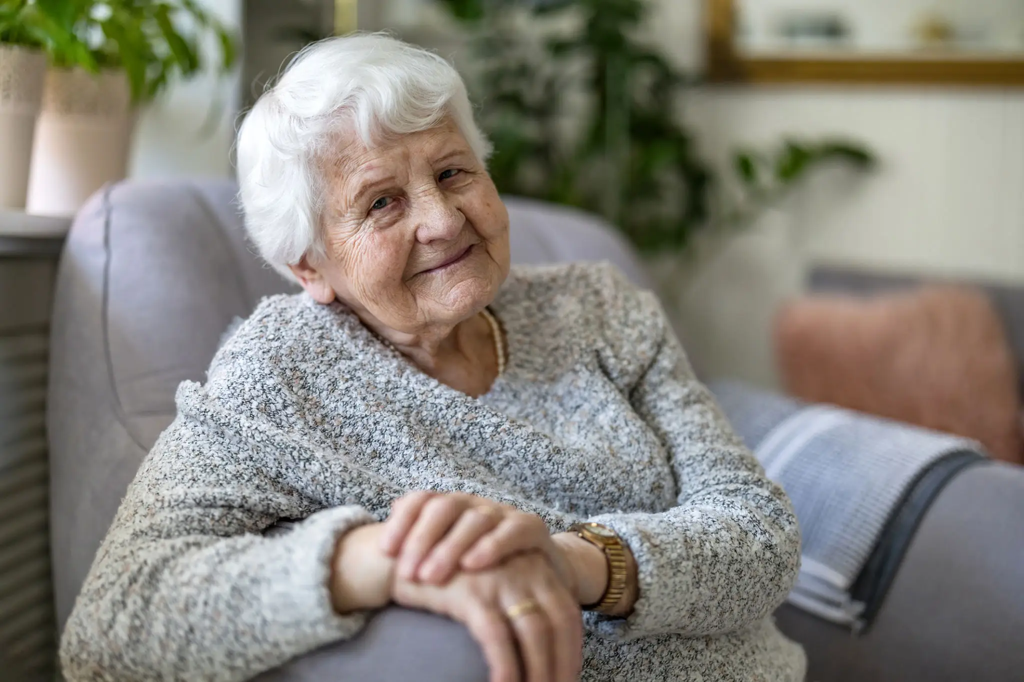 Smiling elderly woman sitting comfortably with hands clasped.